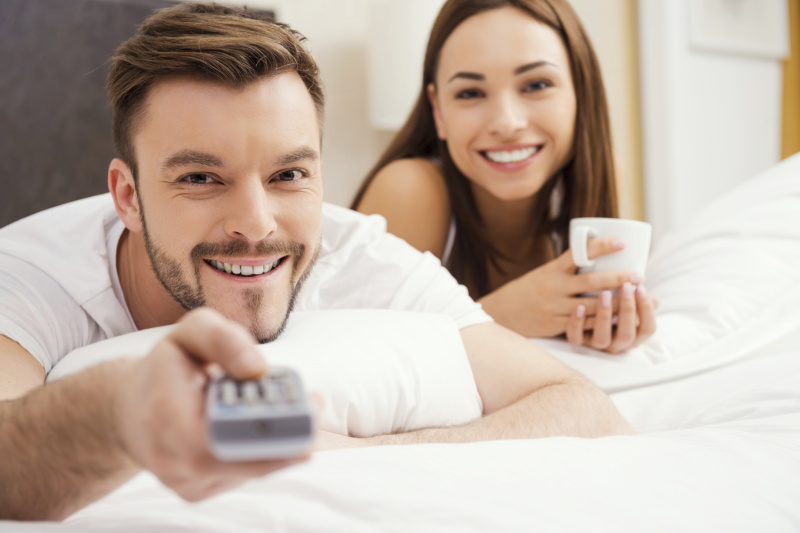 Loving couple in bed. Beautiful young loving couple lying in bed together and smiling while man holding remote control